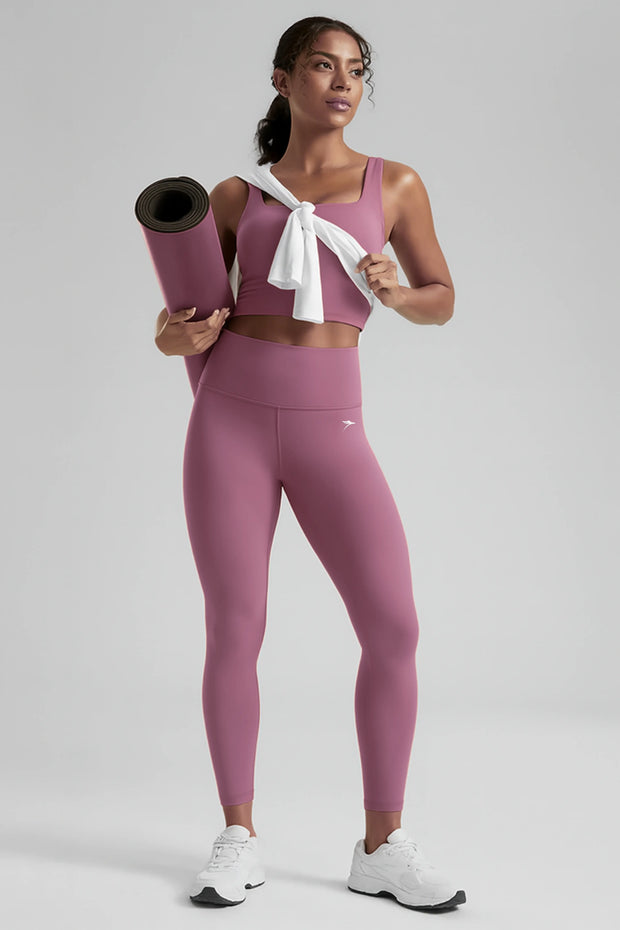 Woman in pink athletic wear holding a yoga mat and towel on a plain background